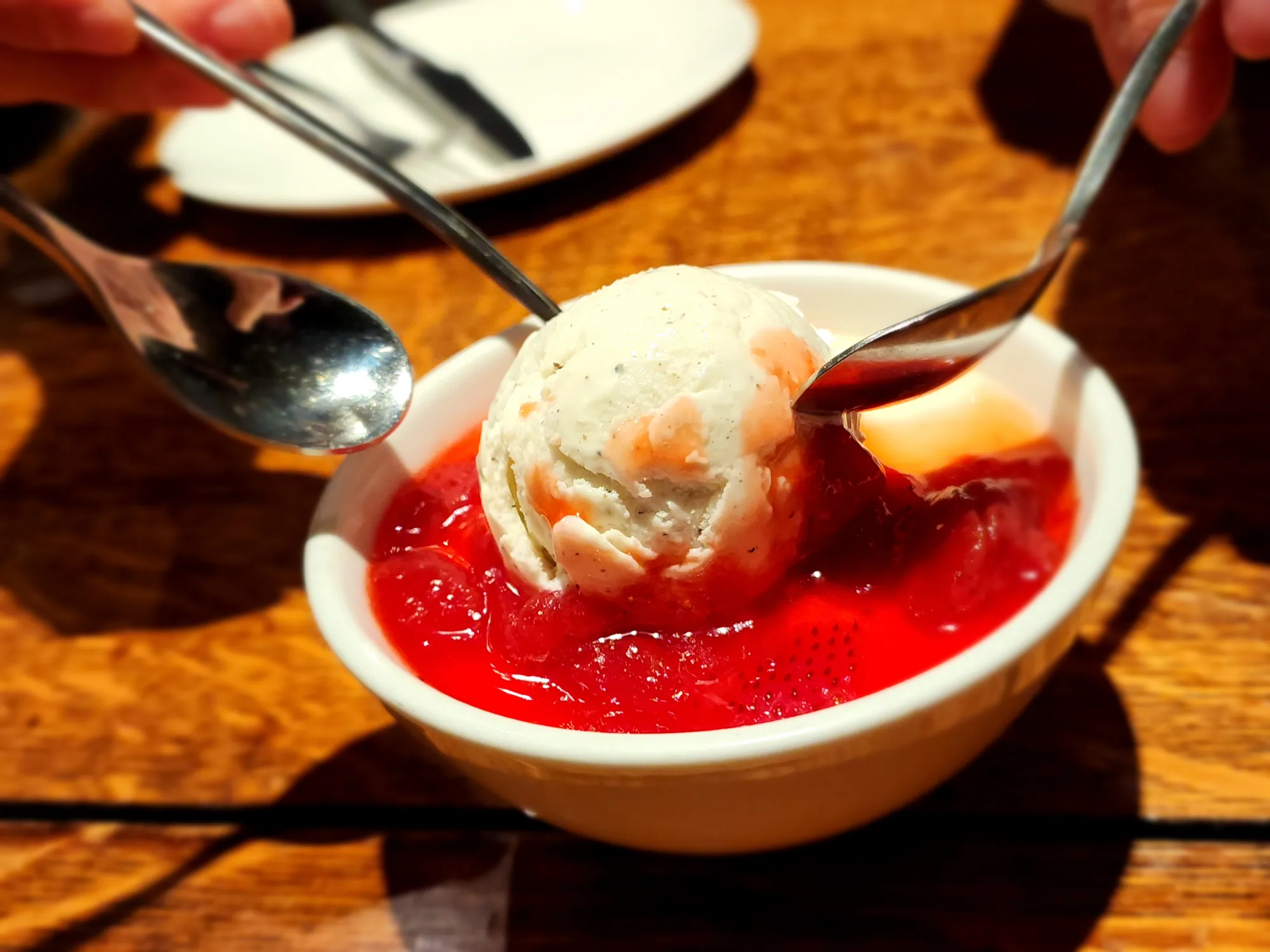A scoop of ice cream over red fruit and sauce, with spoons reaching into the dish