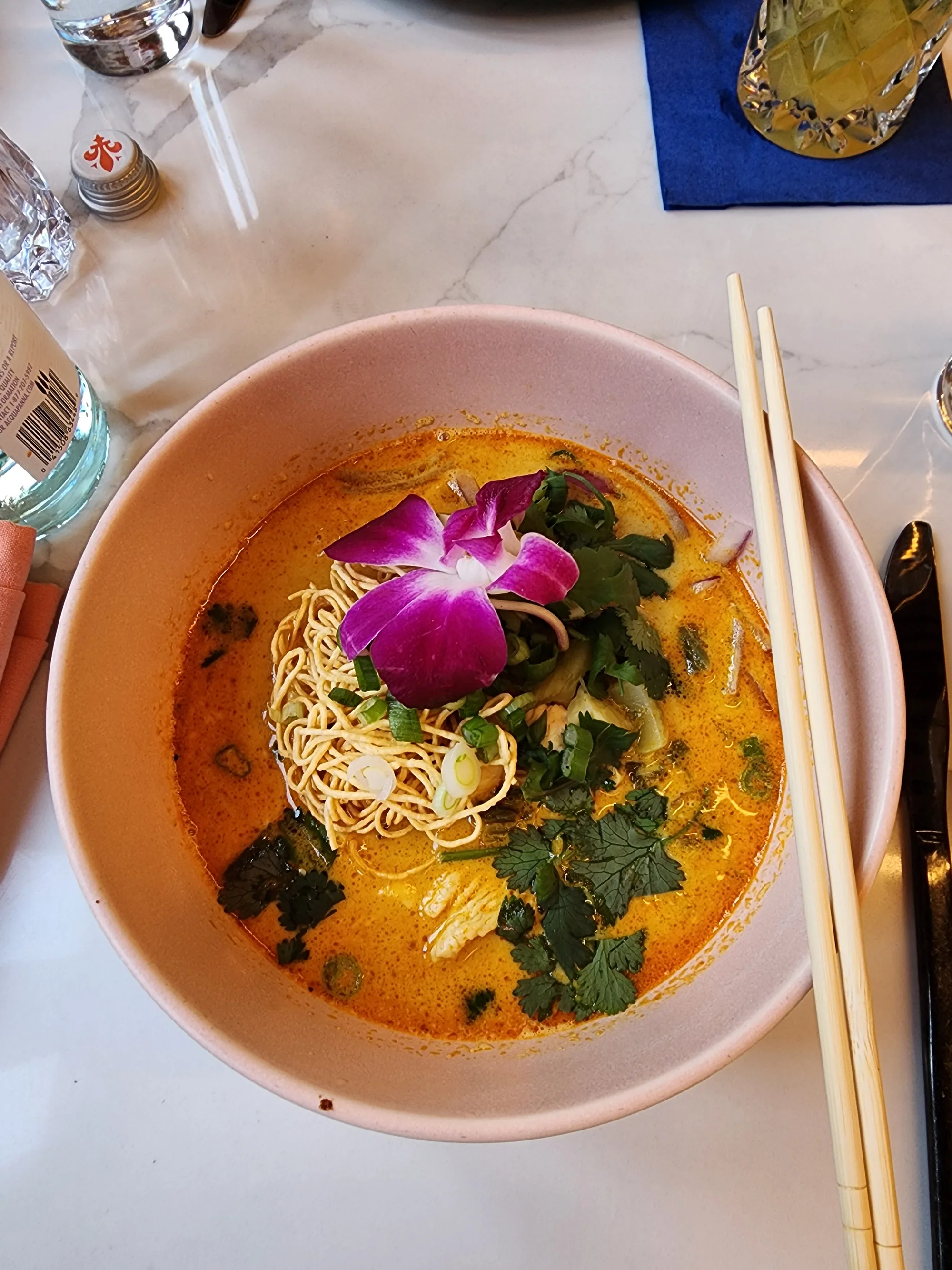 Curry noodles in a pink bowl topped with crisp noodles, herbs, and a purple-and-white flower
