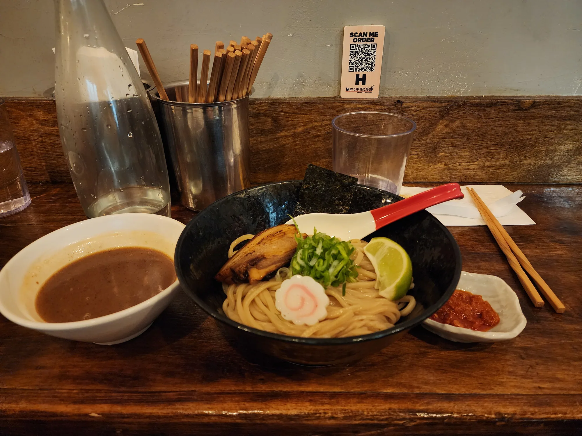 Noodles with pork, scallions, lime, and nori beside separate bowls of dipping sauce and chili sauce on a wooden counter
